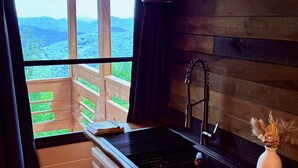 A kitchen with a built-in sink, wooden cabinets, a window with a view of mountains, and a vase with dried flowers. - Black Mtn Stomping Ground's Falcon's Perch with Long-Range Views (Black Mountain)