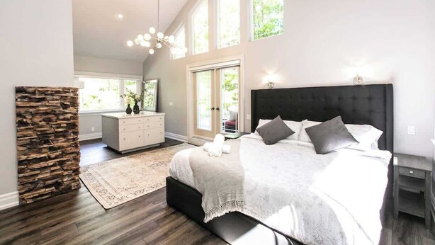A spacious bedroom with a large bed, a stone accent wall, a dresser, and a view of greenery through the windows. - Luxury 4BR Stay with Swim Spa, Golf & Fireplaces (Caledon)