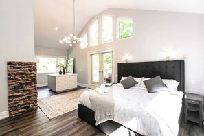 A spacious bedroom with a large bed, a stone accent wall, a dresser, and a view of greenery through the windows. - Luxury 4BR Stay with Swim Spa, Golf & Fireplaces (Caledon)
