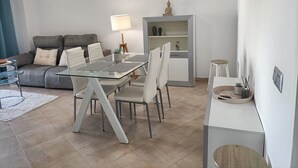 Uma sala de estar moderna com uma mesa de jantar de vidro, cadeiras brancas, um sofá cinzento e uma pequena mesa lateral com uma luminária.