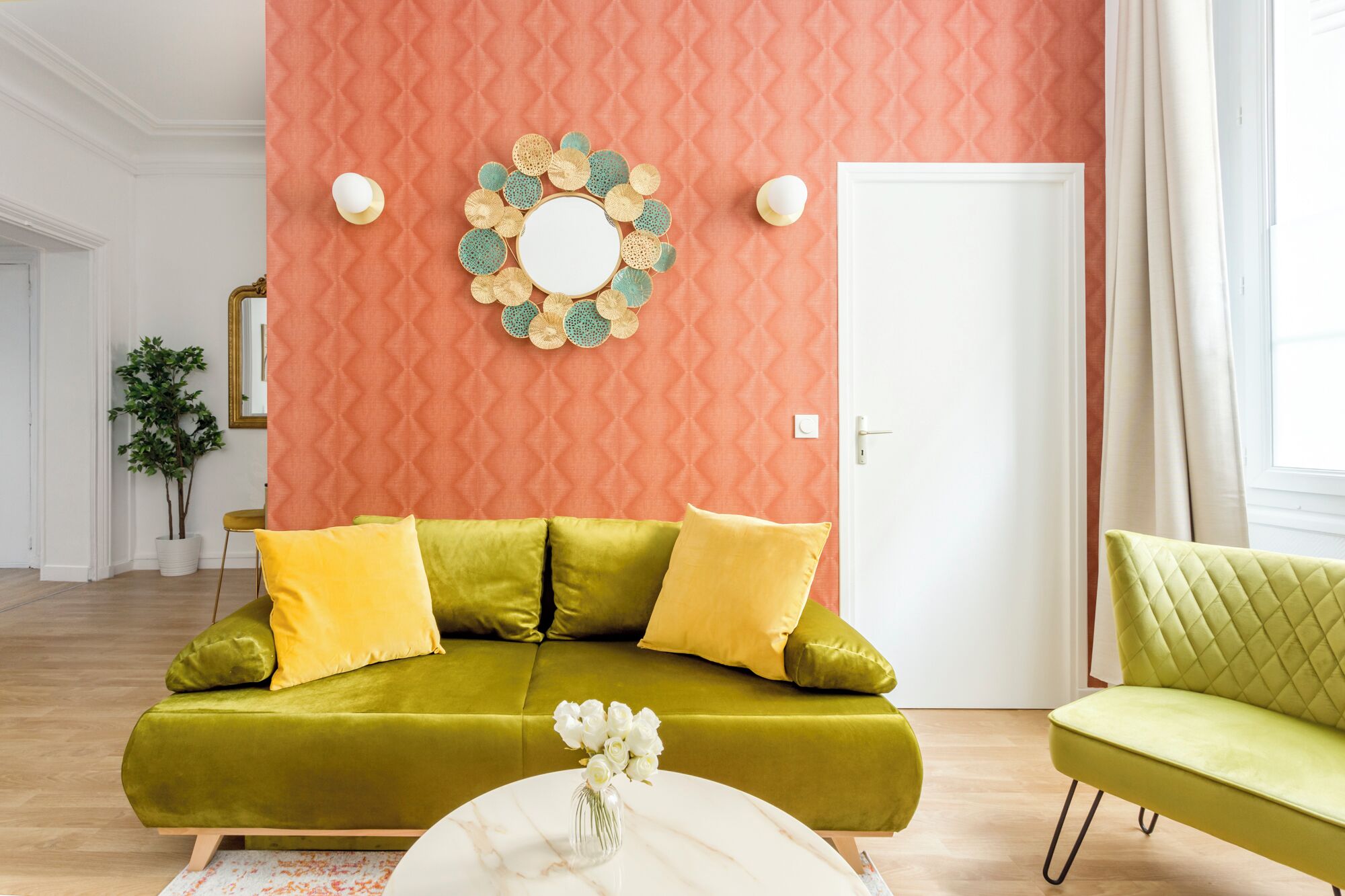 A living room with a green sofa, a green armchair, a round mirror, and a decorative wall hanging.