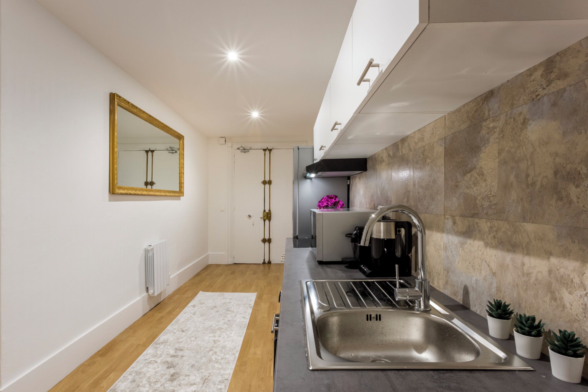 A modern kitchen with a stainless steel sink, a white countertop, and a large mirror on the wall.