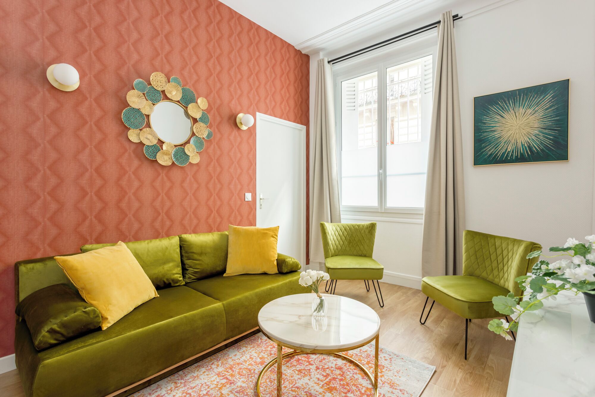 A living room with a green sofa, a round coffee table, and two green armchairs. There is a window with curtains, a mirror with a decorative frame, and a painting on the wall.