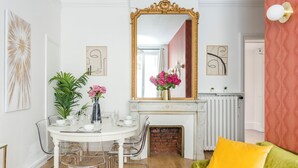 Zona pranzo con tavolo bianco, sedie e un vaso di fiori. Sopra il tavolo, uno specchio con cornice dorata; sotto, un termosifone. In primo piano, un divano verde con un cuscino giallo.