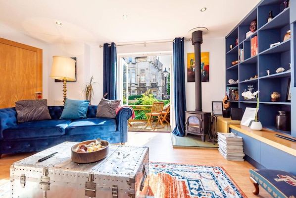 Een woonkamer met een blauwe bank, een vintage koffer, een boekenkast en een houtkachel.