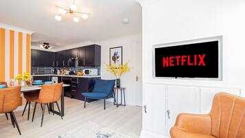 A modern living room with a TV displaying "NETFLIX" and a dining area with orange-striped walls and a kitchen in the background.