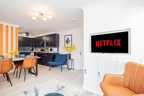 Een moderne woonkamer met een tv waarop “NETFLIX” staat, een eethoek met oranje strepen op de muren en een keuken op de achtergrond.