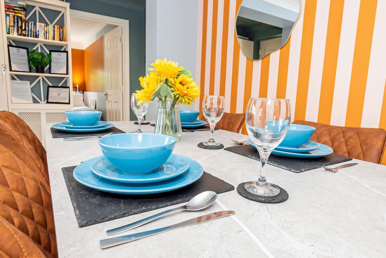 Une table à manger dressée avec des bols, des assiettes et des verres à vin bleus, et un vase rempli de fleurs jaunes placé au centre.