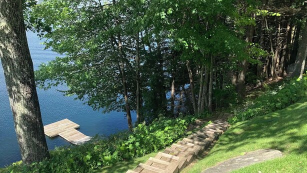 Un escalier en bois descend vers une jetée au bord d’un lac, entouré d’une végétation luxuriante et d’arbres.