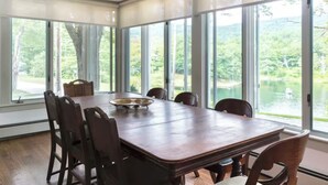 Un coin repas avec une grande table et des chaises en bois, de grandes fenêtres donnant sur un lac et des arbres.