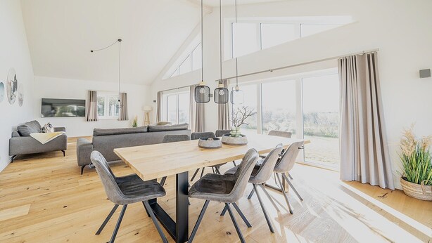 A spacious living area with a wooden dining table, grey sofas, and a large window providing a view of the outdoors. - Ferienhaus in Dagebuell im skandinavischen Stil (Dagebüll)
