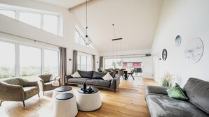 A spacious living room with a large sofa, two armchairs, and a coffee table. There are large windows providing natural light and a view of the outdoors. - Ferienhaus in Dagebuell im skandinavischen Stil (Dagebüll)
