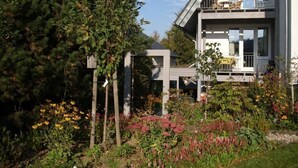Une maison moderne à deux étages, dotée d’un balcon et entourée d’un jardin où poussent diverses plantes et fleurs.