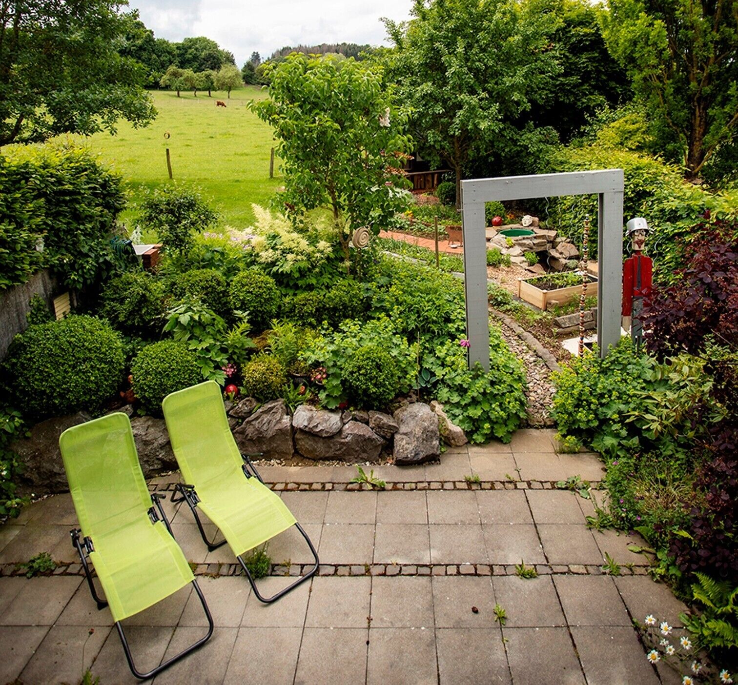 Ein Garten mit einer Metallskulptur, zwei grünen Liegestühlen und einem gepflasterten Weg.