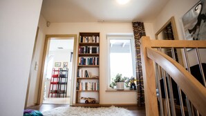 Un couloir avec un escalier en bois, une bibliothèque, une fenêtre avec des rideaux et une porte donnant sur une autre pièce.