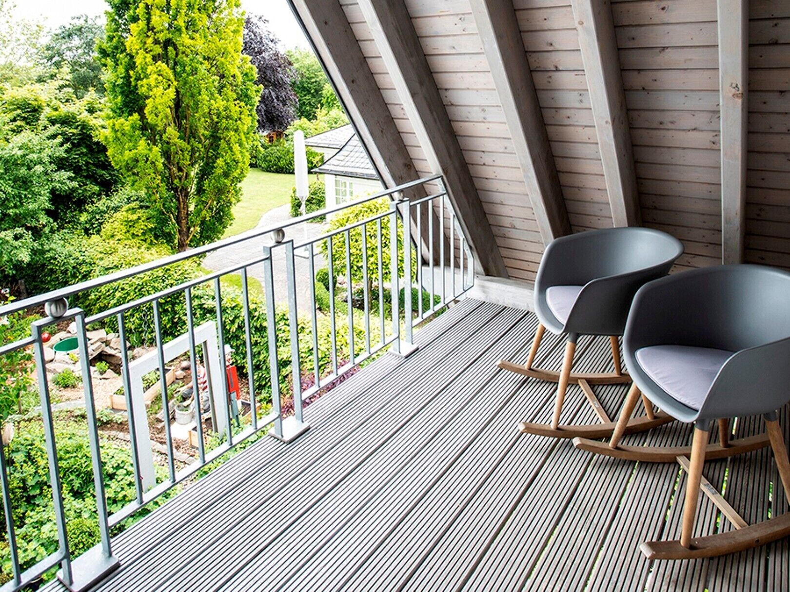 A balcony with a view of a garden, two rocking chairs, and a wooden ceiling.