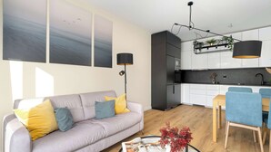 Una sala de estar moderna con un sofá, una mesa de centro y revistas. Hay una zona de comedor con sillas y una mesa. La cocina tiene armarios blancos y una encimera negra.