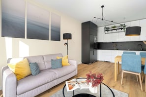 A modern living room with a sofa, coffee table, and magazine. There is a dining area with chairs and a table. The kitchen has white cabinets and a black countertop.
