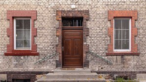 Ein Backsteinhaus mit einer zentralen Holztür, zwei Fenstern mit roten Rahmen und einem kleinen Vorbereich.