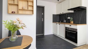 Eine moderne Küche mit weißen Schränken, einer schwarzen Arbeitsplatte und einem eingebauten Ofen. Oberhalb der Spüle befindet sich ein Holzregal und auf dem Tisch steht eine Vase mit Blumen.