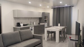 Un salon moderne avec un canapé gris, une table à manger avec des chaises, un coin cuisine équipé d’un four à micro-ondes et une plante en pot.