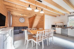 A modern kitchen with a wooden ceiling, a dining area with white chairs, a gray sofa, and a clock on the wall. - Laruns Wooden Cottage (6 Guests) (Laruns)