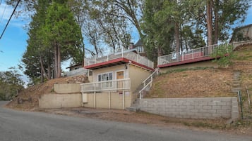 A two-story house with a red roof and white railings, situated on a hillside with trees.