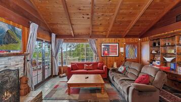 A cozy living room with a fireplace, wooden ceiling, large windows, and a comfortable sofa.
