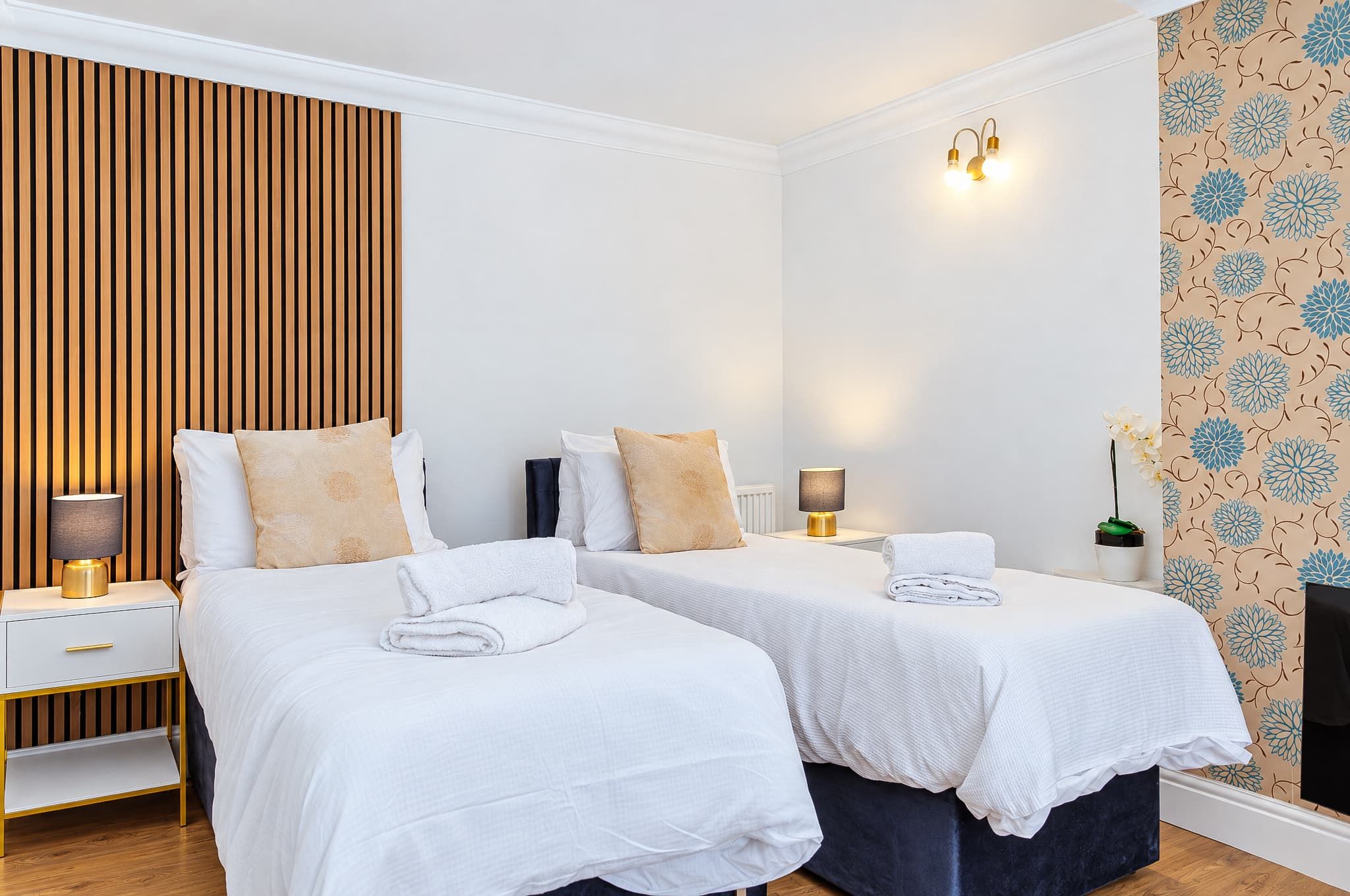 Une chambre d’hôtel avec deux lits, une table de chevet avec une lampe, un luminaire fixé au mur et un papier peint à motifs floraux.