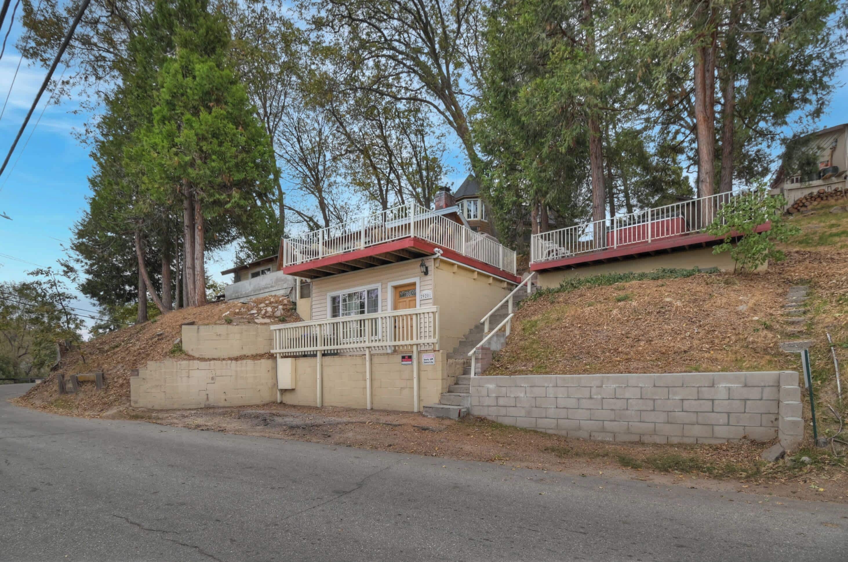 A two-story house with a red roof and white railings, situated on a hillside with trees.