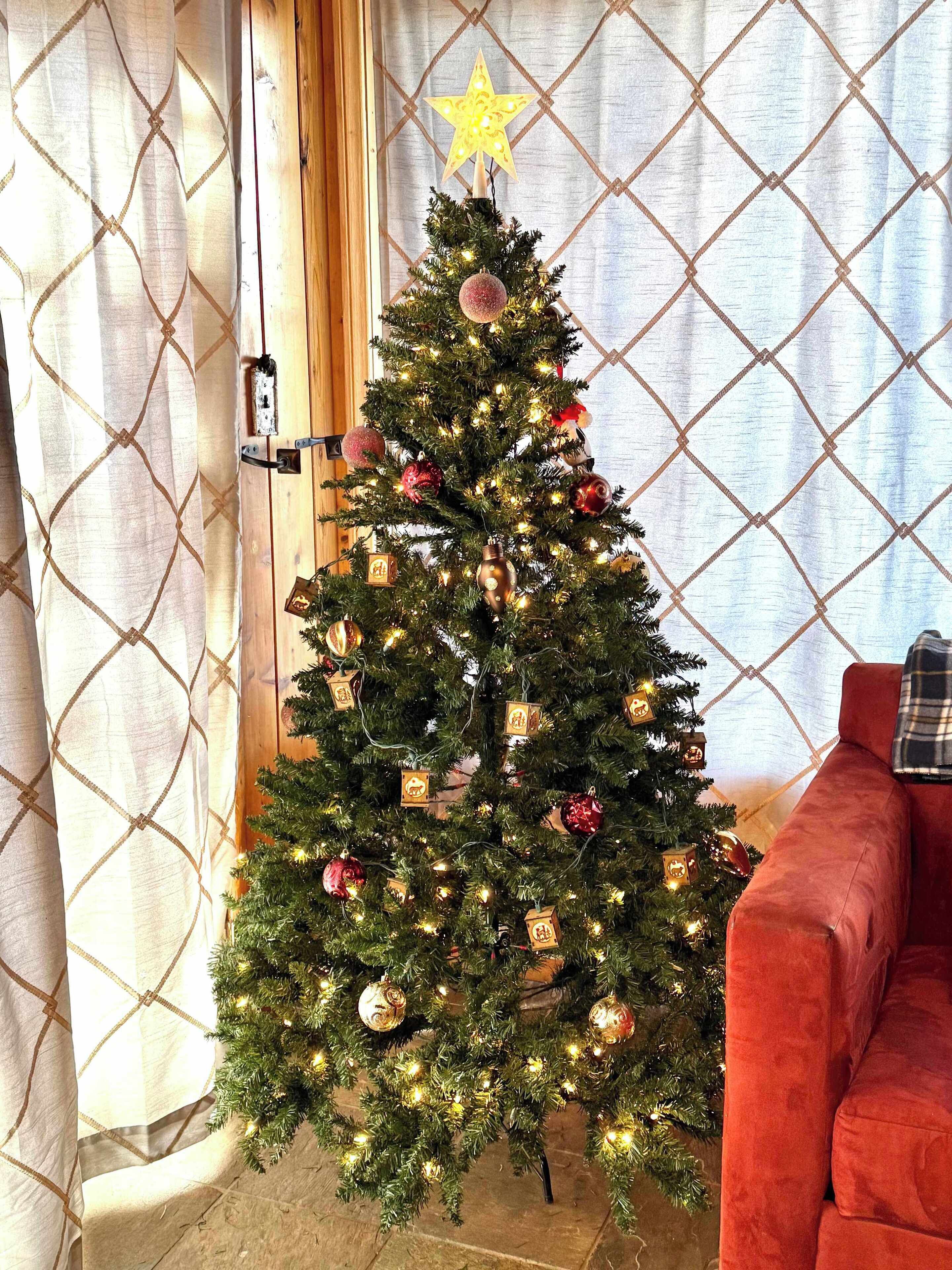 A Christmas tree with ornaments and lights in a room with a red couch and patterned curtains.