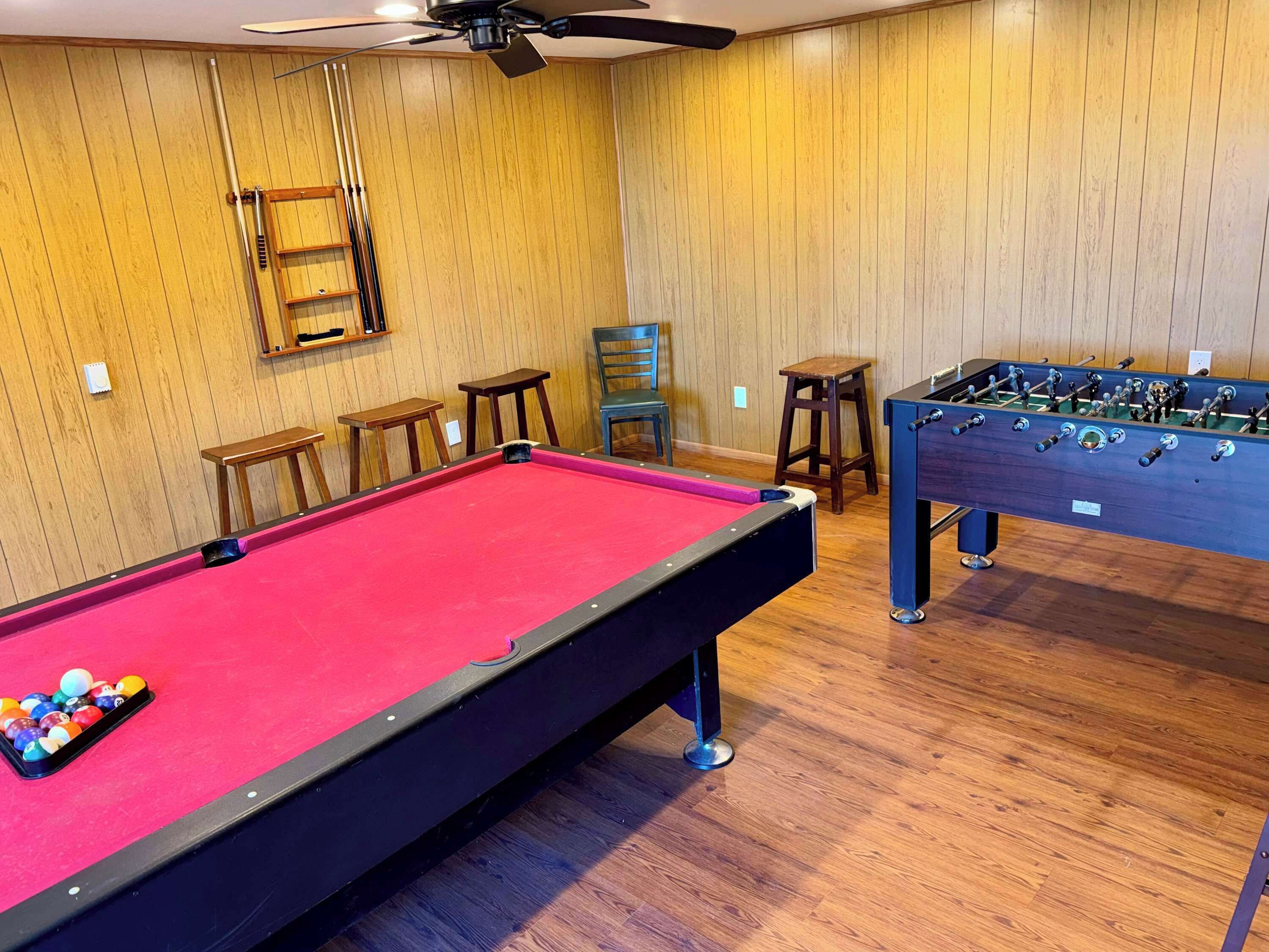 Une table de billard avec des billes, une table de baby-foot et des chaises en bois dans une pièce aux murs en bois et dotée d’un ventilateur de plafond.