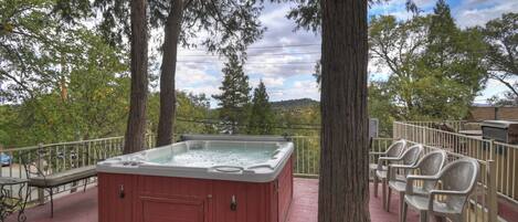 A hot tub on a deck with chairs, surrounded by trees and a view of a hill.