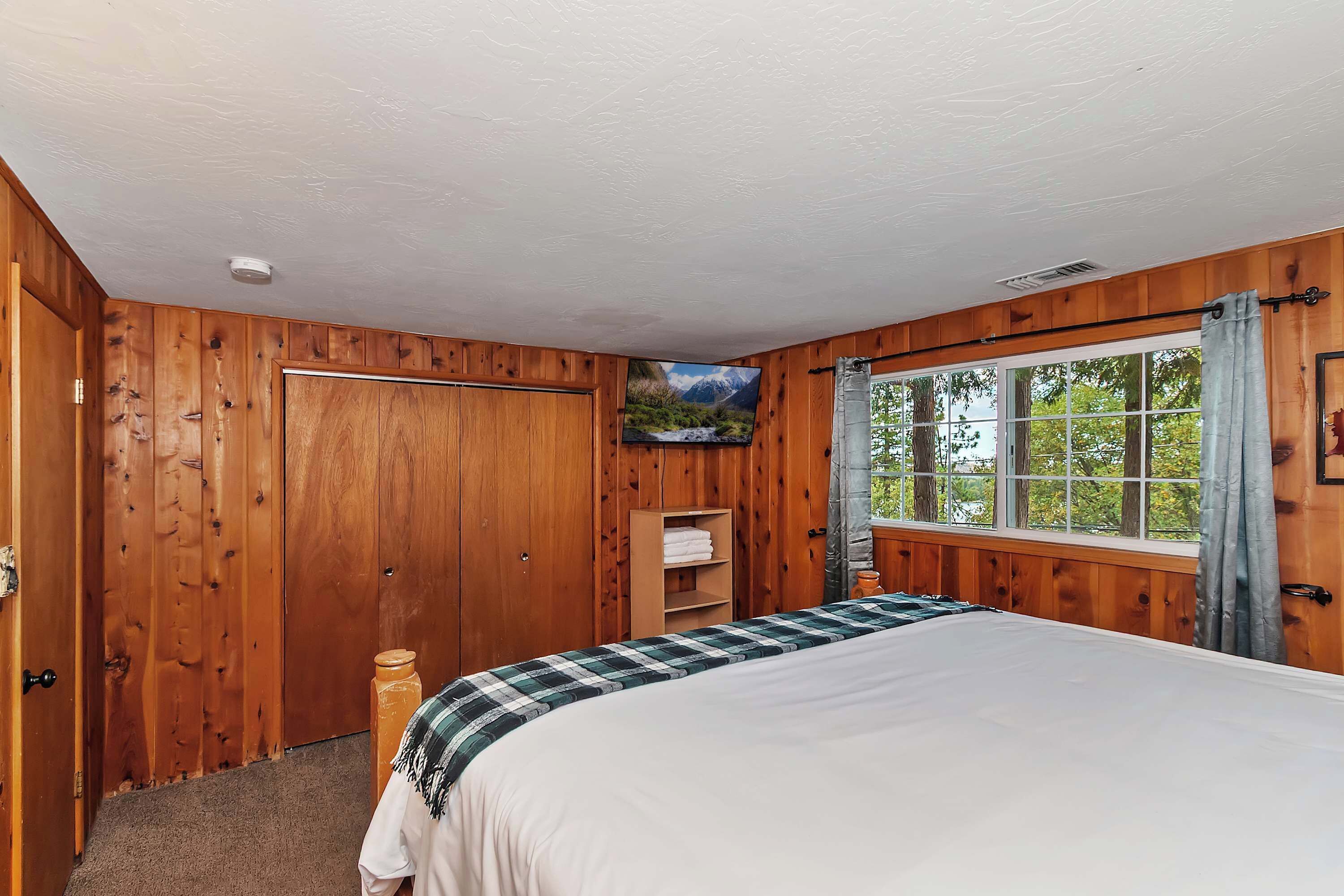 A wooden-paneled bedroom with a bed, a window with curtains, a shelf with towels, and a picture of a mountain.