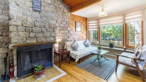 A cozy living room with a stone fireplace, wooden beams, a sofa, and a large window with a view of trees.