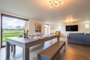 A modern kitchen with a dining table set for four, a bench, and a sofa in a living area with a TV and a painting on the wall.