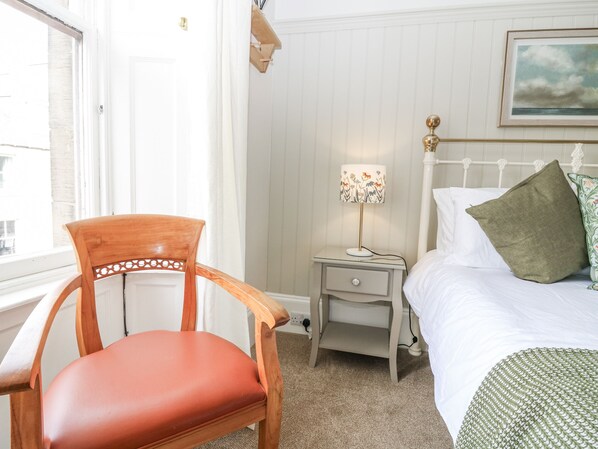 A bedroom with a wooden chair, a bedside table with a lamp, a bed with pillows, and a window with curtains. - 11A High Street (Jedburgh)