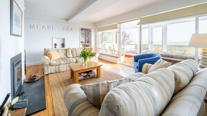 A spacious living room with a large window, a sofa, a coffee table, and a fireplace. - Chicoutimi 4, Thorpeness (Thorpeness)