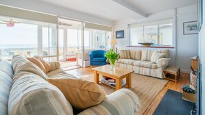A spacious living room with a large sofa, a smaller sofa, a coffee table, and a blue armchair. There is a large window with a view of the sea and a ship in the background. - Chicoutimi 4, Thorpeness (Thorpeness)