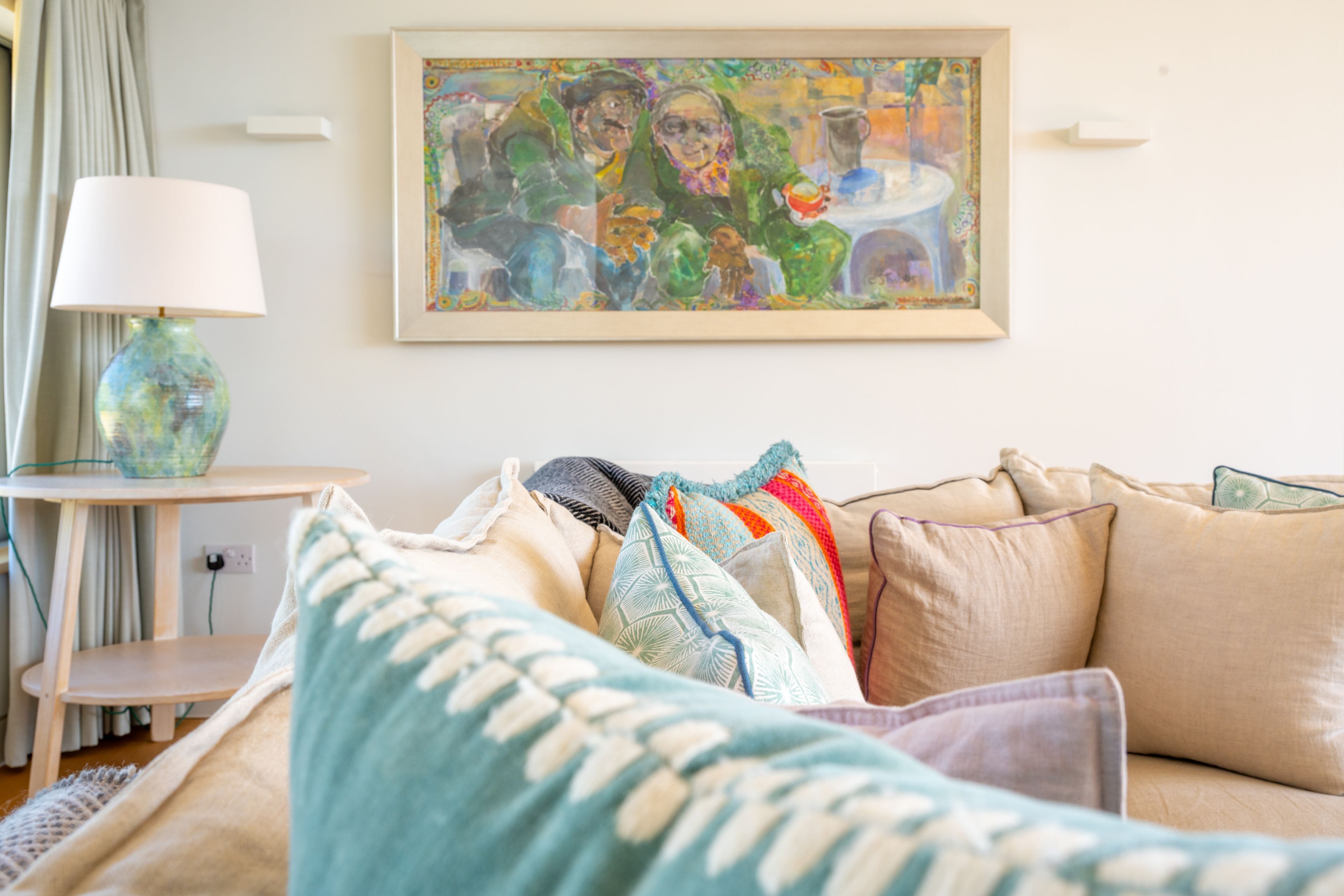 A cozy living room with a beige sofa, patterned cushions, a framed painting, a lamp, and a small table.
