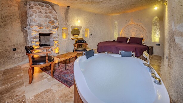 A stone-walled bedroom with a large bathtub, a wooden chair, a small table, and a bed with a red bedspread. - Rose Moon Cave (Nevşehir)