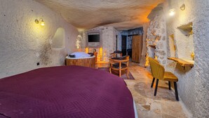 A bedroom with a large bed, a wooden chair, a small table, and a television mounted on a stone wall. - Rose Moon Cave (Nevşehir)