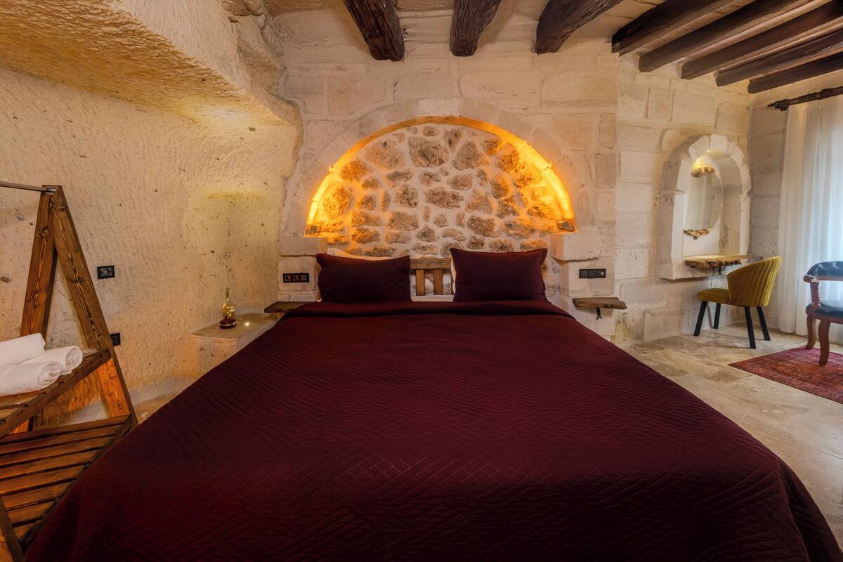 Ein Schlafzimmer mit einem Bogen aus Stein, einer Holzleiter, einem Bett mit roter Decke, einem kleinen Tisch mit Lampe und einem Stuhl.