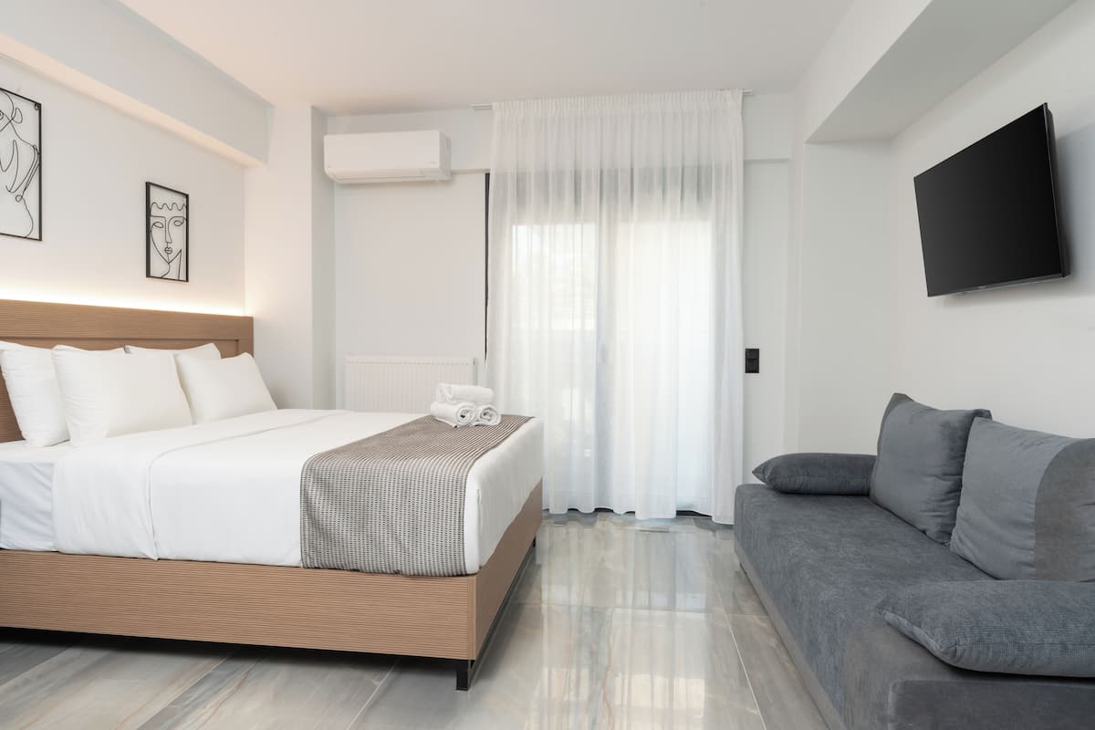 A modern hotel room with a bed, a grey sofa, a flat-screen TV, and artwork on the walls.