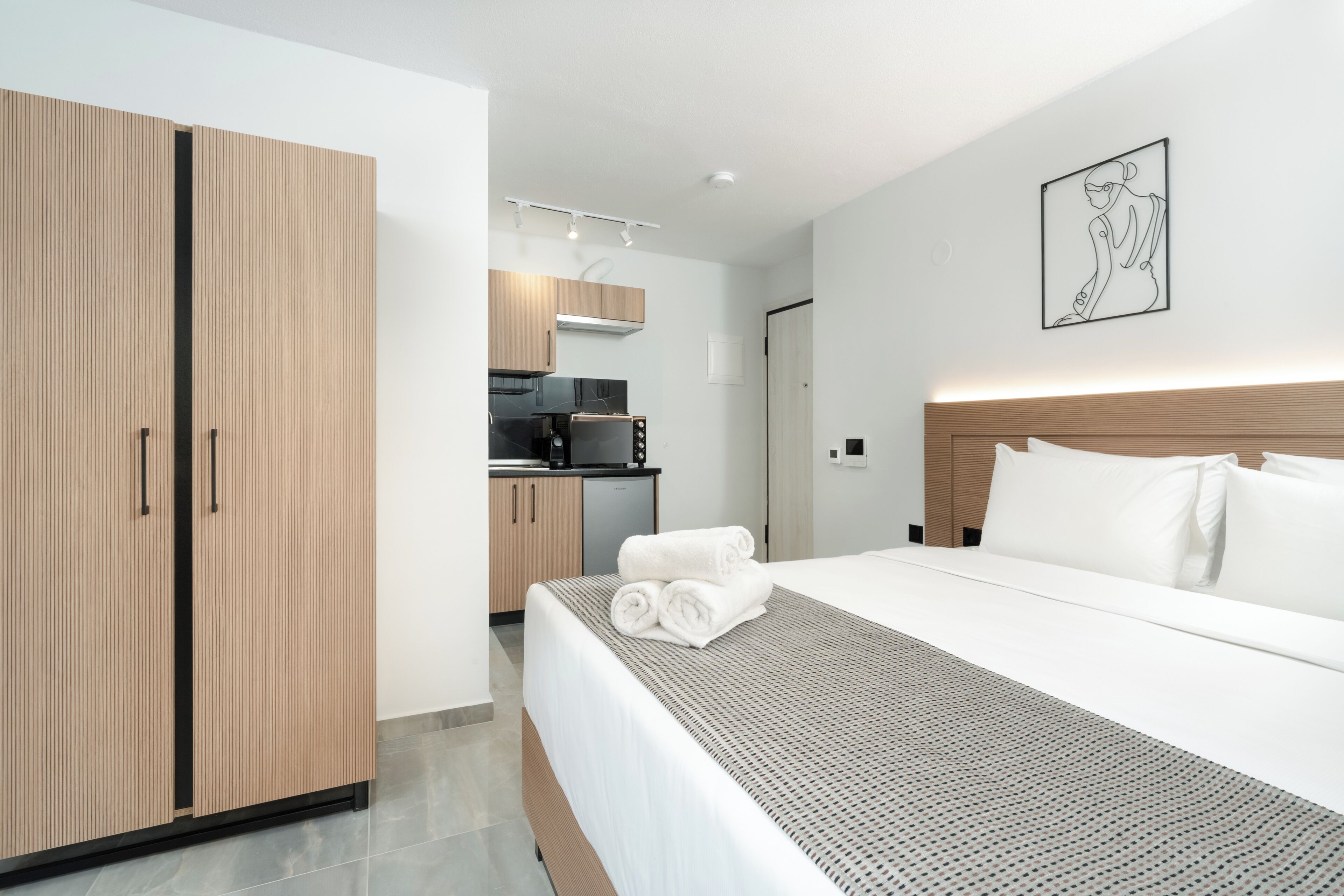 Une chambre d’hôtel moderne avec un grand lit, une armoire en bois, un coin cuisine et une œuvre d’art encadrée au mur.