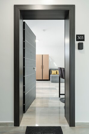 Un interior moderno con una puerta de madera oscura, suelo de baldosas de color claro, un armario de madera, un sofá gris con un cojín amarillo y un dispositivo negro fijado a la pared.