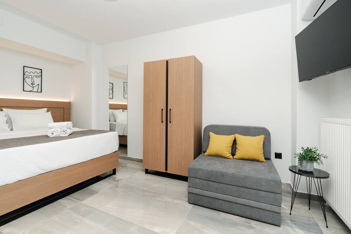 A modern bedroom with a bed, a grey sofa, a wooden wardrobe, and a small table with a plant.