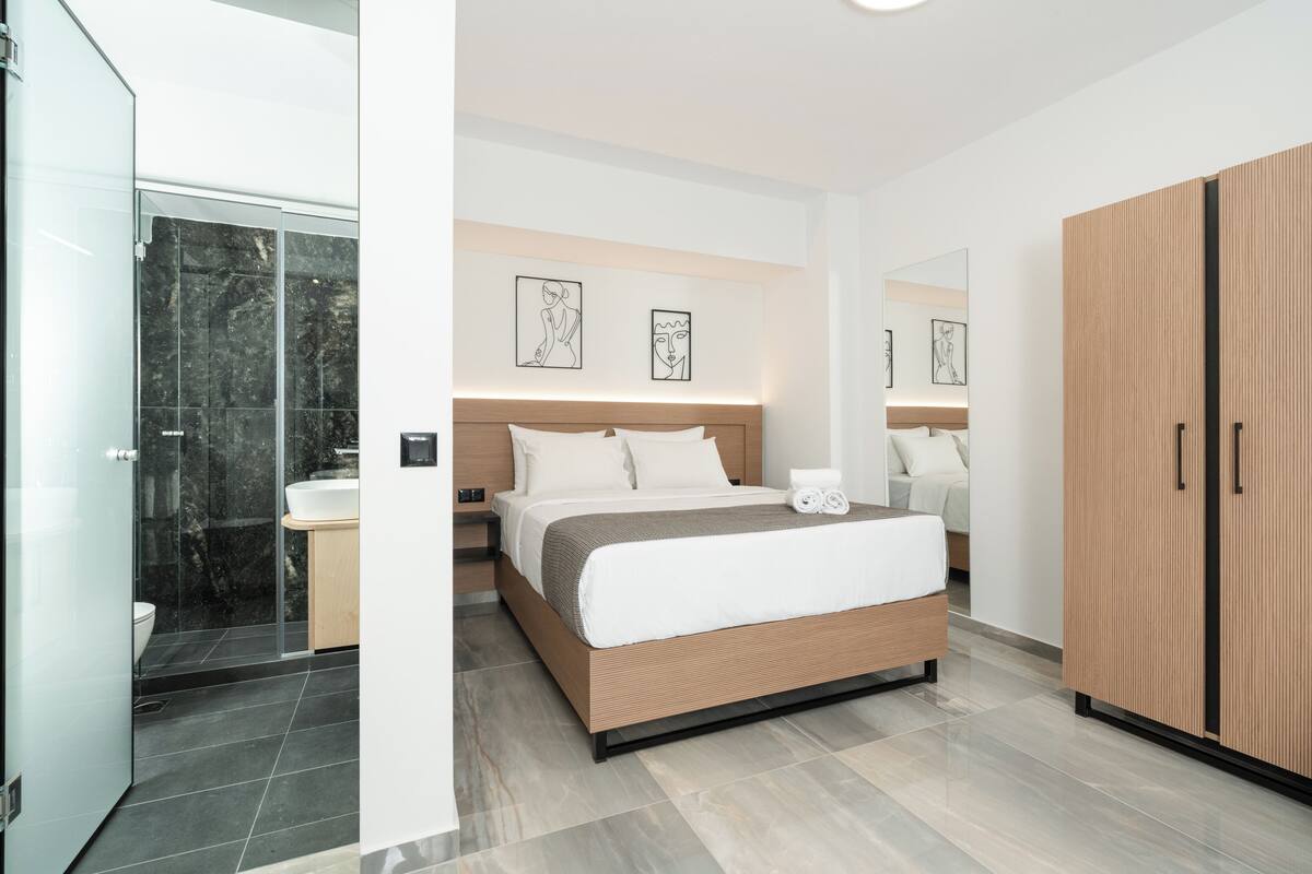 A modern hotel room with a large bed, a wooden wardrobe, a bathroom with a glass shower enclosure, and artwork on the walls.