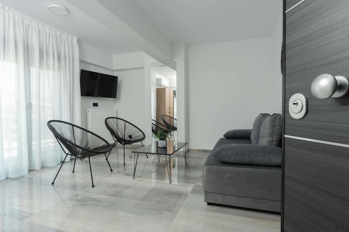 A modern living room with a grey sofa, two black wireframe chairs, a glass coffee table, and a flat-screen TV mounted on the wall.