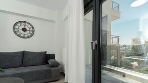 A modern living room with a grey sofa, a round wall clock, and a view of the city through a glass door. - TOWER 15 Suites (Thessaloniki)